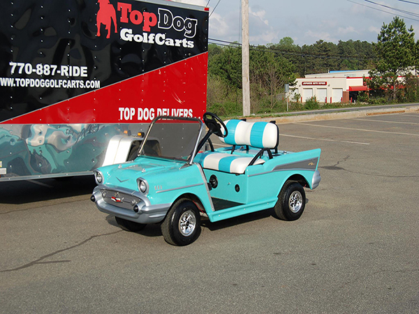 A turquoise and white retro-style golf cart is parked on pavement near a Top Dog Golf Carts trailer.