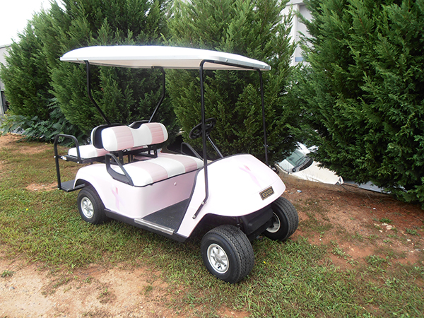 A pastel pink and white golf cart with a roof is parked on grass beside tall green bushes.