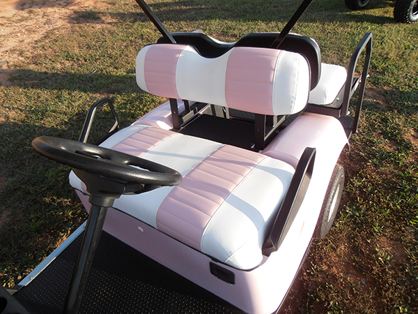 A pink and white golf cart with two bench seats is parked on grass in sunlight.