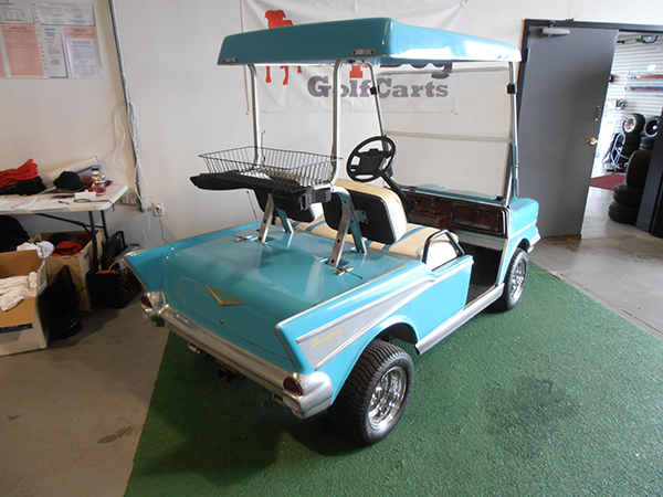 A blue golf cart with a retro car design, featuring a basket on the front and white seats, is parked indoors on green turf near an open door.