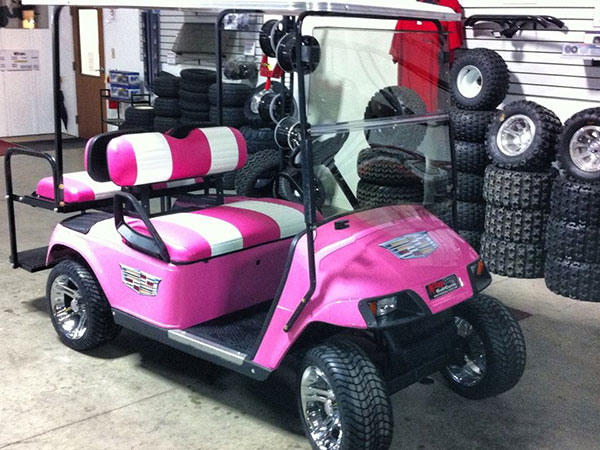 A pink and white golf cart with custom seats is parked indoors next to stacks of spare tires and wheels.