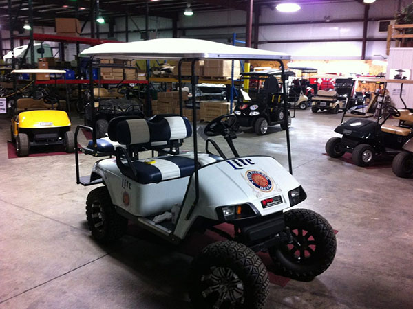 A custom white golf cart with off-road tires is parked inside a warehouse filled with other golf carts and boxes.