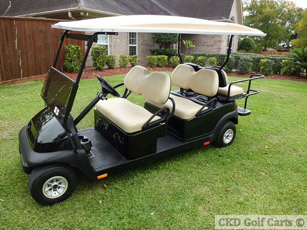 A black four-seater golf cart with beige seats is parked on a grassy lawn near a house and wooden fence.
