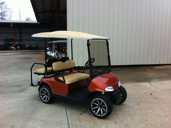 A red and beige golf cart with black wheels is parked on a concrete surface near a white metal building.