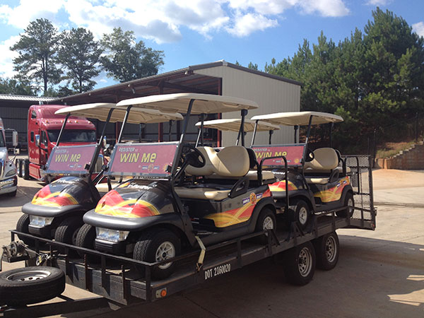 Three golf carts with "WIN ME" signs are loaded on a flatbed trailer parked outside a building, with trees and trucks in the background.