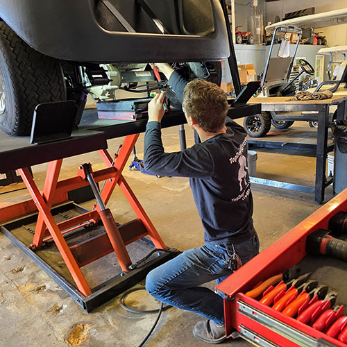 A mechanic kneels beside a lifted golf cart, servicing its underside in a workshop with tools and equipment visible nearby.