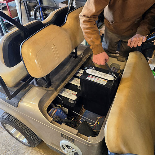 Person performing maintenance on the battery compartment of a beige golf cart, with seat lifted to access the batteries and tools in hand.