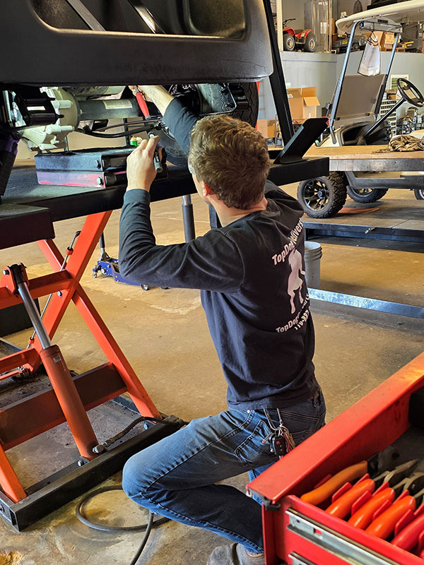 A mechanic kneels on the floor while working underneath a lifted golf cart inside a workshop, with tools and equipment visible nearby.