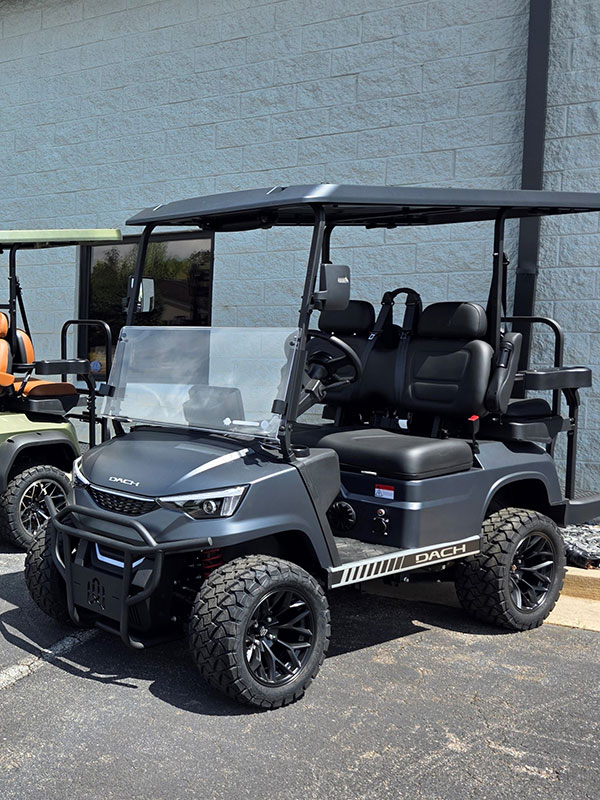 A black, four-seater golf cart with rugged tires is parked on asphalt near a light gray building. Another green golf cart is partially visible beside it.