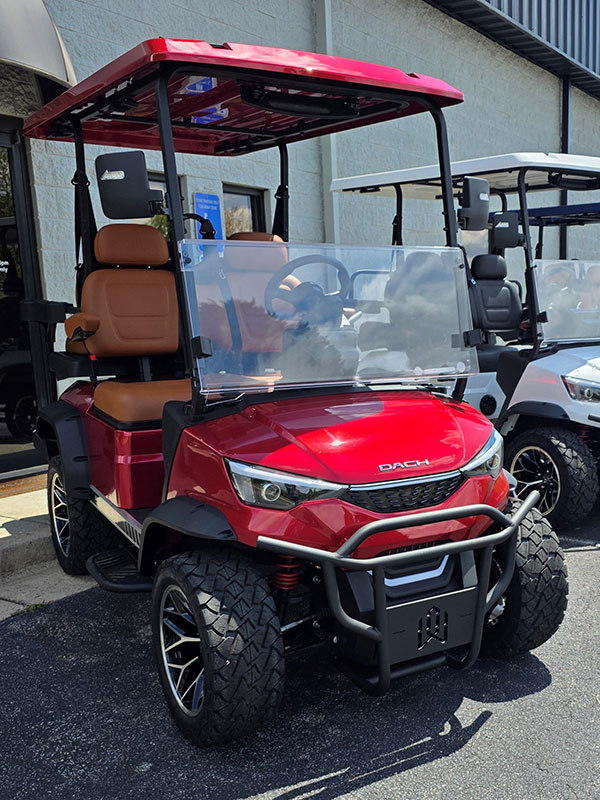 A red golf cart with brown seats and a clear windshield is parked outside a building next to another golf cart.