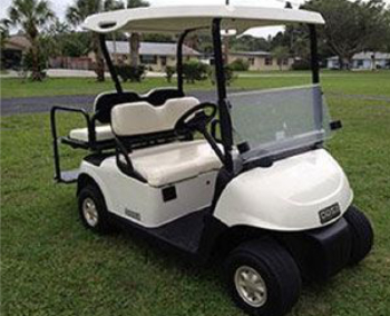 A white golf cart with black trim and beige seats is parked on grass in an outdoor area with houses and palm trees in the background.